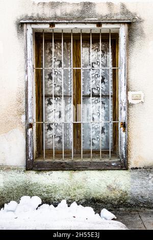 Altes Holzfenster mit Eisenstangen und Vorhängen, Schnee auf dem Boden Stockfoto