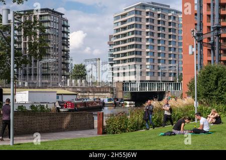 Die Menschen sitzen auf Gras und gehen den Weg, Regent’s Canal am King’s Cross, London. Schmale Boote, hohe Gebäude und Oberleitungen im Hintergrund. Stockfoto