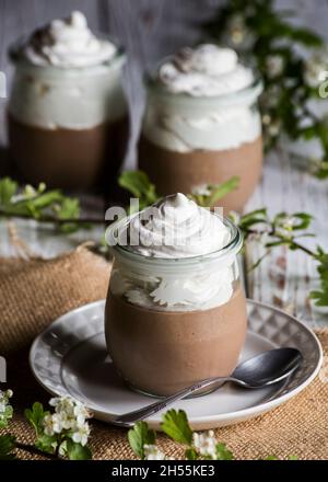 Eine Schokoladenmousse mit Cremecreme. Selektiver Fokus. Glasbehälter auf einem weißen Holztisch mit einem Stück hessischen Tuch und einem Zweig wi dekoriert Stockfoto