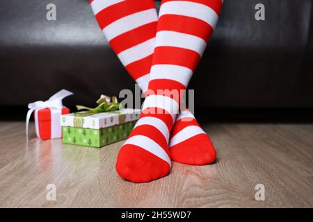 Weibliche Beine in Weihnachtskniesocken auf einem Boden in der Nähe der Geschenkkartons. Frau, die zu Hause auf einem Bett sitzt, Neujahrsfeier Stockfoto