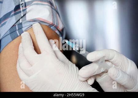 Impfung während der Covid-19-Pandemie, weibliche Hand in Schutzhandschuh mit Spritze. Krankenschwester bereitet sich auf die Injektion in eine männliche Schulter vor Stockfoto