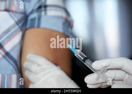 Impfung während der Covid-19-Pandemie, weibliche Hand in Schutzhandschuh mit Spritze. Krankenschwester bereitet sich auf die Injektion in eine männliche Schulter vor Stockfoto