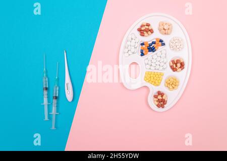 Globale Pharmaindustrie und Medizinprodukte - verschiedenfarbige Pillen, Tabletten und Kapseln auf White Art Palette mit Spritze und Thermometer auf Split Blue und Pink Hintergrund Stockfoto