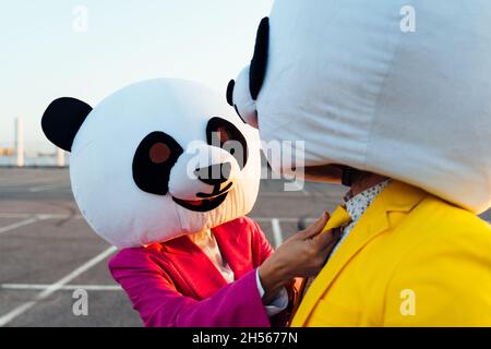 Ein erzähltes Bild eines Paares, das einen riesigen Pandakopf und farbige Anzüge trägt. Mann und Frau feiern auf einem Parkplatz. Stockfoto