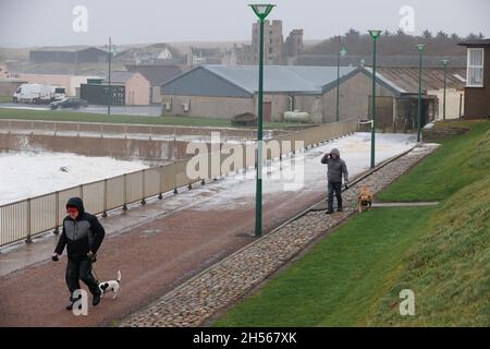 Thurso, Schottland. Nov. 7 2021. Zwei Männer laufen mit ihren Hunden am Wasser entlang, während die Windböen in Thurso, Caithness, Schottland, stürmen. Stockfoto