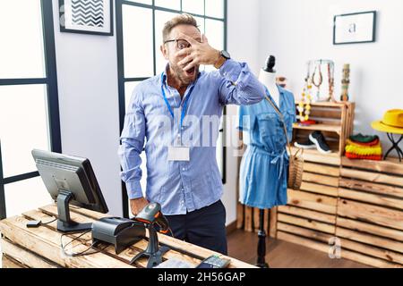 Mann mittleren Alters, der als Manager in einer Einzelhandelsboutique arbeitet und mit der Hand ins Gesicht und in die Augen schauend, mit beschämter Anspannung durch die Finger schauend Stockfoto