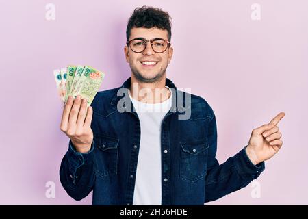 Junger hispanischer Mann mit argentinischen Pesos-Banknoten lächelnd und mit Hand und Finger zur Seite zeigend Stockfoto