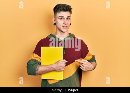 Junge kaukasische Junge mit Ohren Erweiterung halten Buch lächelnd glücklich zeigt mit Hand und Finger Stockfoto