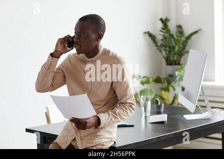 Waist-up-Porträt eines schwarzen Geschäftsmannes, der mit dem Smartphone am Schreibtisch spricht, in minimalem Innenraum, Platz zum Kopieren Stockfoto