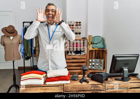 hispanischer Mann mittleren Alters, der als Manager in der Einzelhandelsboutique arbeitet, lächelt fröhlich und guckst einen Buh mit Händen, die Gesicht zeigen. Überrascht und begeistert Stockfoto