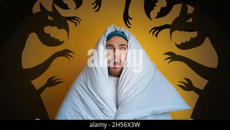 Verängstigender junger Mann, der sich vor seinen inneren Ängsten versteckt, eingewickelt in eine Decke auf orangefarbenem Hintergrund mit Monstersilhouetten Stockfoto