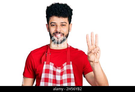 Junger arabischer Mann mit Bart und Kochschürze, der mit den Fingern Nummer drei nach oben zeigt und dabei selbstbewusst und glücklich lächelt. Stockfoto