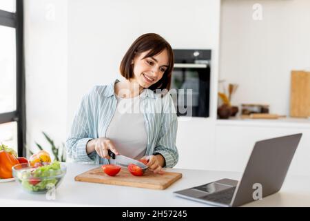 Junge Frau kocht frischen Bio-Salat, schneidet Tomaten und schaut auf Laptop auf der Arbeitsplatte, versucht neues Rezept Stockfoto