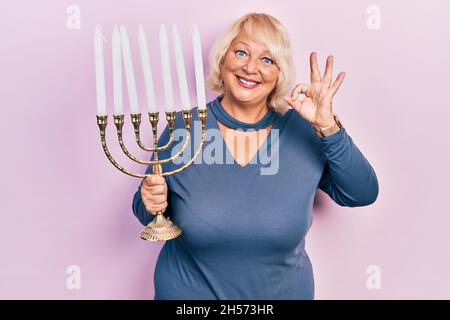 Mittelalte blonde Frau hält Menorah hanukkah jüdische Kerze tun ok Zeichen mit den Fingern, lächelnd freundlich gestikulieren ausgezeichnetes Symbol Stockfoto