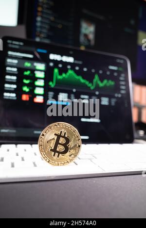 Eine Bitcoin-Münze steht auf einer Computertastatur und im Hintergrund befindet sich das Bitcoin-Diagramm Stockfoto