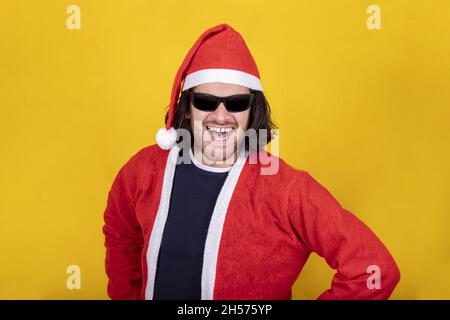 Glücklicher und attraktiver Mann ist als Weihnachtsmann gekleidet. Er trägt eine Sonnenbrille und einen roten Hut. Gelber Hintergrund und Platz zum Kopieren Ihrer Nachricht. Stockfoto