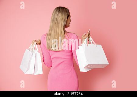 Nicht erkennbare schlanke blonde Frau mit langen Haaren Rückansicht hält Papier Einkaufstaschen mit Käufen Studio auf rosa Hintergrund geschossen, shopaholic Stockfoto