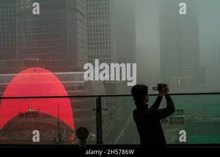 Ein junger Mann fotografiert einen Wolkenkratzer, der in starkem Smog gehüllt ist, im Geschäftszentrum von Peking, China. 06-Nov-2021 Stockfoto