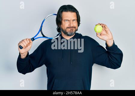 Kaukasischer Mann mittleren Alters spielt Tennis mit Schläger und Ball skeptisch und nervös, runzelte die Stirn wegen des Problems. Negative Person. Stockfoto