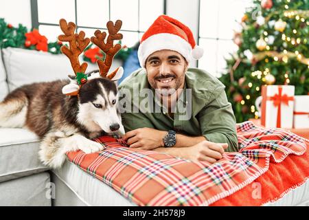 Junger hispanischer Mann, der zu Hause auf dem Sofa liegt und einen Hund trägt, der einen Rentierhut vom Weihnachtsbaum trägt Stockfoto