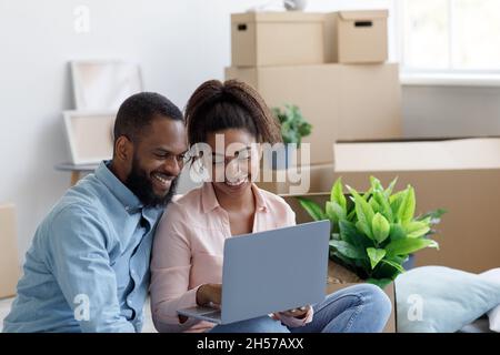 Lächelnder junger schwarzer Mann und Dame in einer neuen Wohnung zwischen Boxen, Planung der Innenausstattung, Paar mit Laptop Stockfoto
