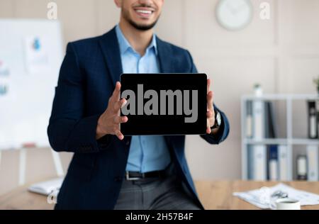 Nicht erkennbarer arabischer Geschäftsmann, der einen Tablet-Computer mit leerem Bildschirm zeigt und ein Modell für eine Website oder eine App im Büro anbietet Stockfoto