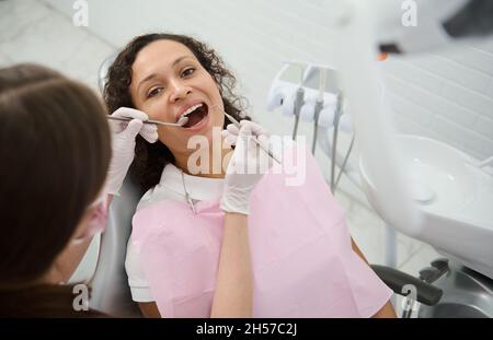 Die junge Patientin mit einem schönen Lächeln sitzt mit offenem Mund auf dem Zahnarztstuhl, während die Hygienistin ihre Zähne mit Stainl untersucht oder behandelt Stockfoto