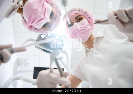 Bodenansicht eines Zahnarztes und eines Zahnarztes in rosa Schutzmaske, die zahnärztliche Werkzeuge und Instrumente zur Untersuchung hält und sich zur Patie neigt Stockfoto