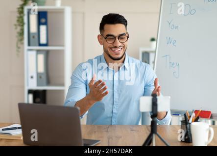Fernlernkonzept. Fröhliche arabische Lehrerin, die online Englischunterricht gibt, mit Smartphone und Laptop im Heimbüro Stockfoto