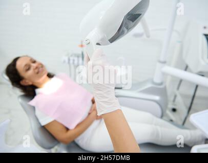 Nahaufnahme der Hand eines Zahnarztes in einem chirurgischen Handschuh, der das Licht gegen einen verschwommenen lächelnden Patienten anpasst, der auf dem Zahnarztstuhl sitzt und auf de wartet Stockfoto