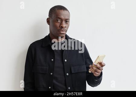 Minimal Taille up Porträt von schwarzen jungen Mann hält Smartphone und Blick auf die Kamera, während an der weißen Wand stehen, kopieren Raum Stockfoto