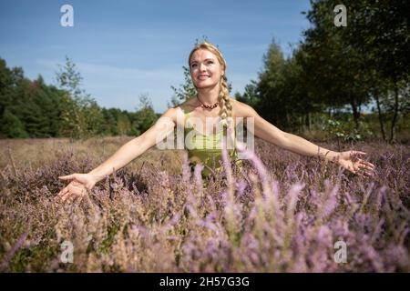 Eine schöne junge Kräuterkundige breitet ihre Arme weit aus und umarmt die Heideblumen. Feldkräuter. Stockfoto