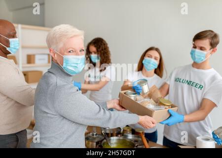 Humanitäre Hilfe. Gruppe junger Freiwilliger mit Gesichtsmasken, die älteren Menschen Boxen mit Produkten und Konserven geben Stockfoto