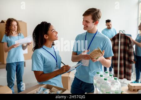 Junge schwarze Frau überprüft die Liste der Wohltätigkeitsorganisationen, während die Gruppe od Freiwillige Lebensmittelpakete für Menschen in Not anstellt Stockfoto