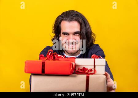 Ein langhaariger junger Mann verteilt Weihnachtsgeschenke, Pakete mit rotem, braunem Papier und rotem Band. Gelber Hintergrund und Kopierbereich. Stockfoto