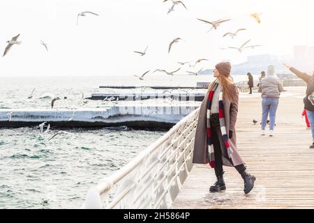 Wanderfrau auf dem Pier im Winter. Stilvolle Dame trägt Wintermantel, raue Stiefel, Schal und Hut allein in der Nähe des Meeres stehen Stockfoto