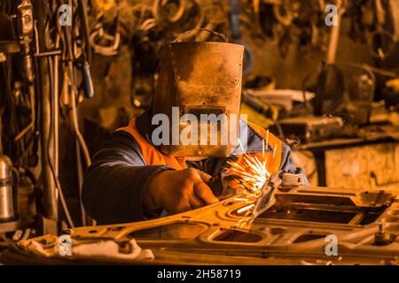 Der Schweißer in einer eisernen Schutzmaske repariert die Autotür und ist in der Werkstatt des Industriewerks mit Schweißarbeiten beschäftigt. Stockfoto