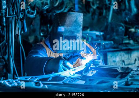 Ein männlicher Schweißer aus dem Baugewerbe in einem Schutzschild ist mit Schweißarbeiten an Metall und der Reparatur einer Autotür in der Werkshalle beschäftigt. Stockfoto