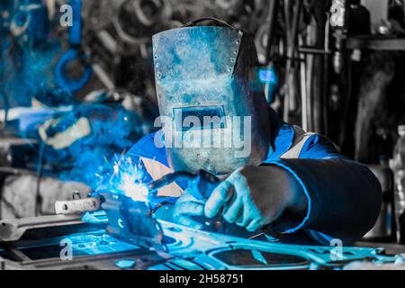 Der Schweißermann im Schutzschild beschäftigt sich mit der Schweißen- und Metallbearbeitung, der Reparatur der Autotür in der Werkstatt des Werks. Stockfoto