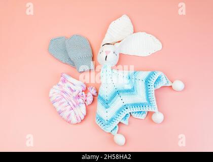 Handgefertigte gestrickte pink-weiße Stiefelette, kleine graue Fäustlinge, blaues und weißes Hase-Spielzeug für Frühgeborene, isoliert auf rosa Hintergrund Stockfoto