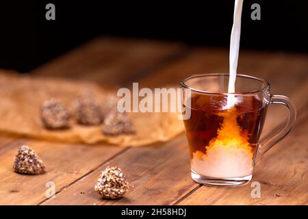 Ein Strom Milch fließt in eine Tasse Tee. Tasse auf einem hölzernen Hintergrund um Pralinen. Stockfoto