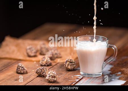 Ein Milchstrahl strömt in eine Tasse Tee, die einen Spritzer bildet. Milch wird auf einem Holztisch verschüttet. Süßigkeiten auf dem Tisch. Stockfoto