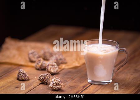 Ein Strom Milch fließt in eine Tasse Tee. Tasse auf einem hölzernen Hintergrund um Pralinen. Stockfoto