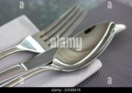 Metallsuppenlöffel, Messer, Gabel liegen auf einer grauen Serviette auf einer schwarz-grauen Oberfläche Draufsicht. Tischeinrichtung, Tuch im Restaurant, Café, in der heimischen Küche. Grau Stockfoto