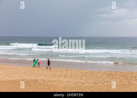 Zwei Surfer standen am Manly Beach, als ein Mann in Schwarz an einem bewölkten Tag in Sydney vorbeiwanderte, Australien Stockfoto