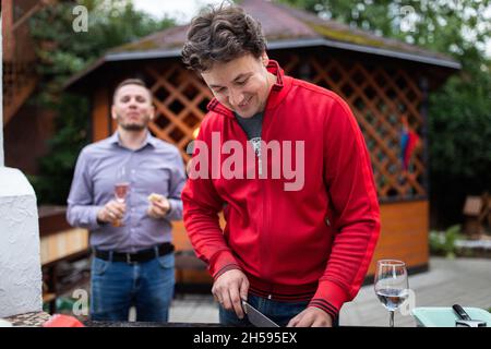 Ein Mann bereitet auf einer Hinterhofparty Essen für seine Freunde zu. Er schneidet Pizzaprodukte, im Hintergrund trinkt ein Freund Champagner. Stockfoto