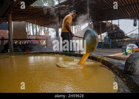 Roorkee, Indien. November 2021. Ein Arbeiter gießt dicken Zuckerrohrsaft in einen anderen Behälter, während er Zange (traditionelle Zuckerrohrkonfektion) herstellt.Zange die Herstellung aus Zuckerrohrsaft ist in vielen Teilen Indiens eine traditionelle ländliche Industrie. Mehr als 70 % der weltweiten Produktion von Jaggery werden von Indien produziert. Kredit: SOPA Images Limited/Alamy Live Nachrichten Stockfoto