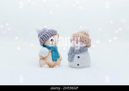 Dekorative Figuren im Weihnachtsstil. Die Statuette einer Katze und eines Schneemanns in einem gestrickten Hut auf weißem Hintergrund. Festliche Einrichtung, warme Bokeh-Lichter Stockfoto