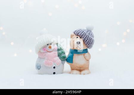 Dekorative Figuren im Weihnachtsstil. Die Statuette eines Bären und eines Schneemanns in einem gestrickten Hut auf weißem Hintergrund. Festliche Einrichtung, warmes Bokeh-Licht Stockfoto