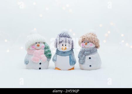 Dekorative Figuren im Weihnachtsstil. Die Statuette eines Pinguins und zweier Schneemänner in einem gestrickten Hut auf weißem Hintergrund. Festliche Einrichtung, warmes Bokeh Stockfoto